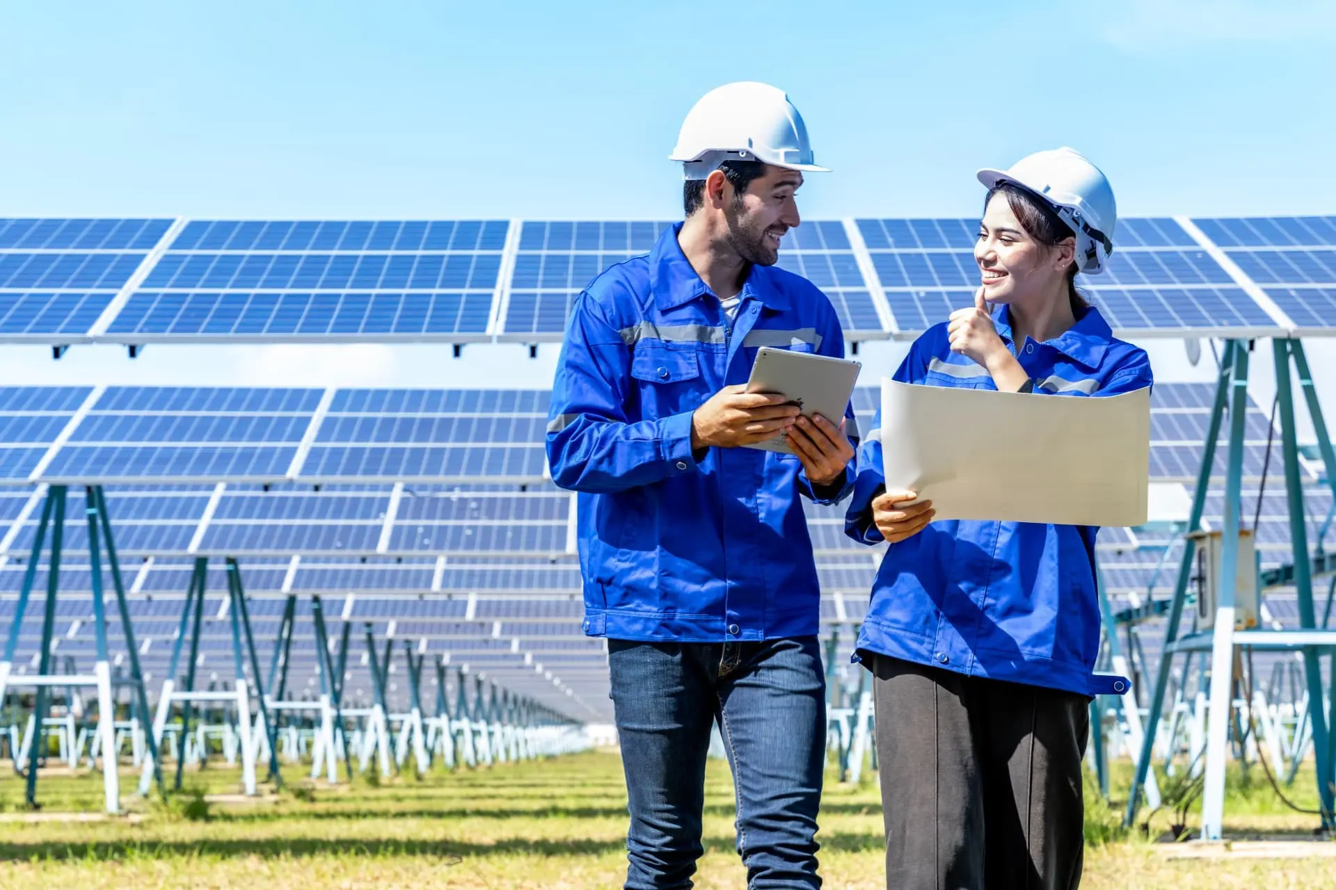 Um engenheiro e uma engenheira elétrica, ambos vestindo capacetes de segurança brancos e uniformes de trabalho azuis, estão em uma usina de energia solar. Eles sorriem enquanto revisam um projeto em papel e um tablet digital, com painéis solares fotovoltaicos estendendo-se ao fundo. A imagem sugere a inspeção e o planejamento de um sistema solar digital.