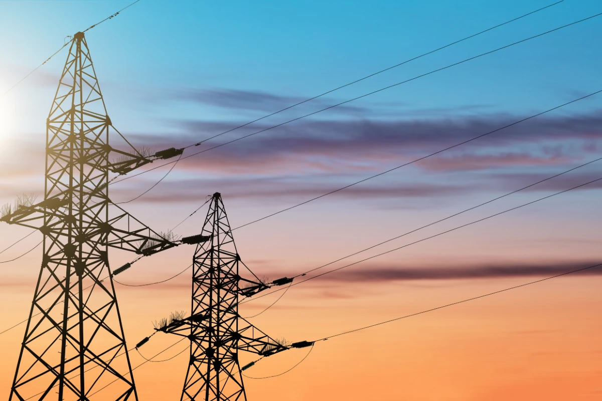 Silhuetas de duas torres de transmissão de alta tensão (linhas de energia) contra um céu em tons de azul e laranja-avermelhado ao pôr do sol. Os cabos de transmissão se estendem pela imagem.