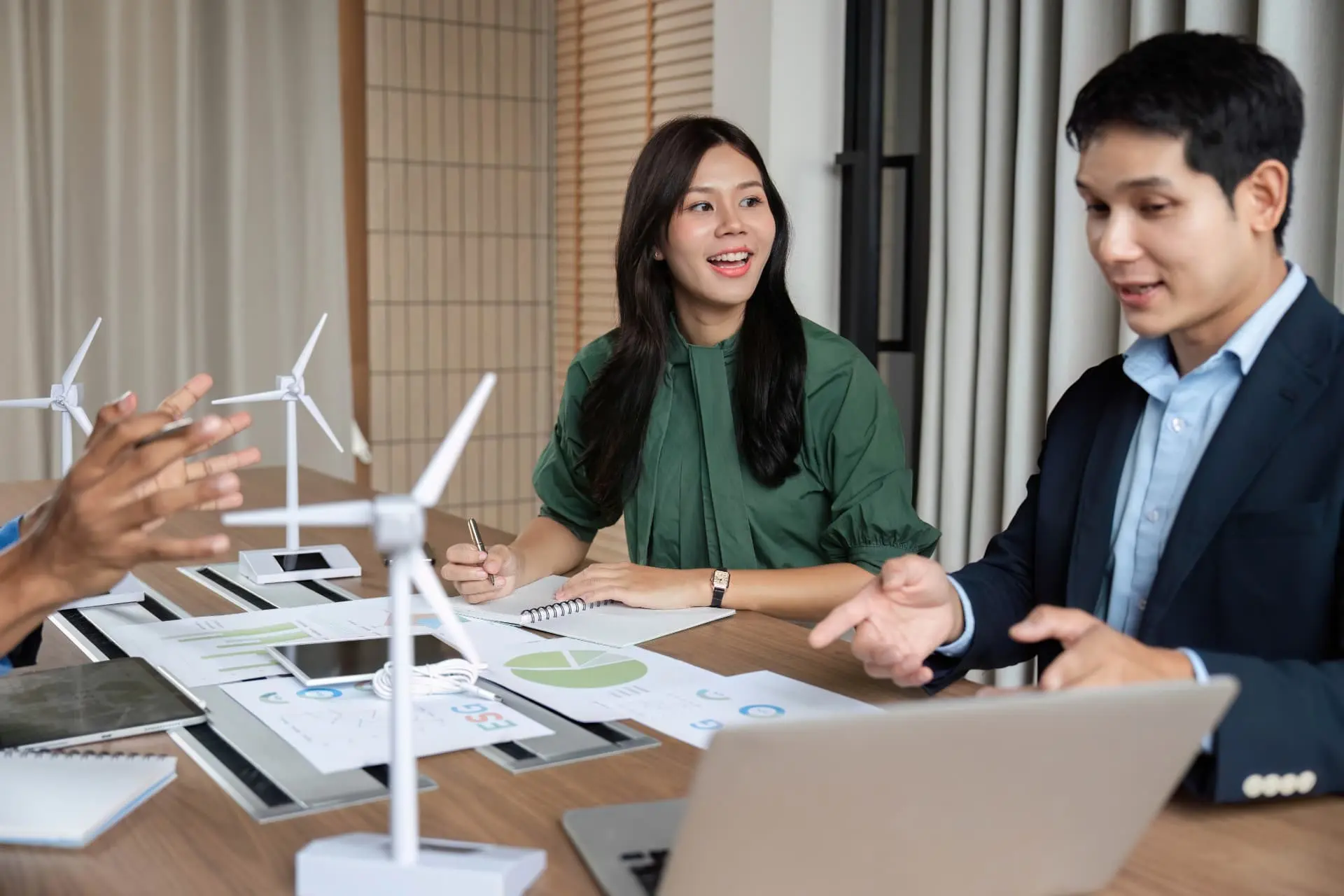 Três profissionais (duas mulheres e um homem, supostamente engenheiros ou consultores) debatem em torno de uma mesa com vários modelos de turbinas eólicas e documentos de análise financeira (gráficos e a sigla ESG). O homem à direita gesticula e explica, enquanto a mulher no centro sorri e anota, evidenciando uma discussão estratégica sobre energia sustentável e sustentabilidade corporativa.