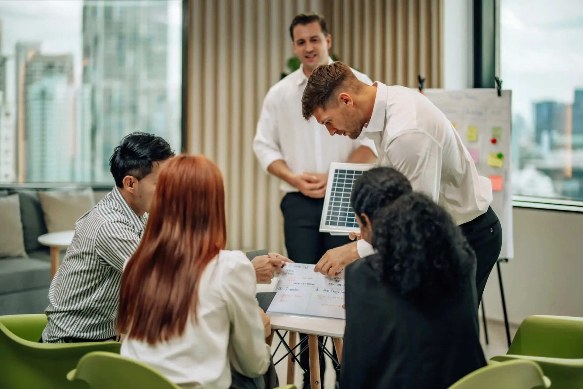 Quatro empresários em uma reunião em escritório moderno, com vista para a cidade. Um homem de camisa branca se inclina e apresenta um projeto em uma prancheta.